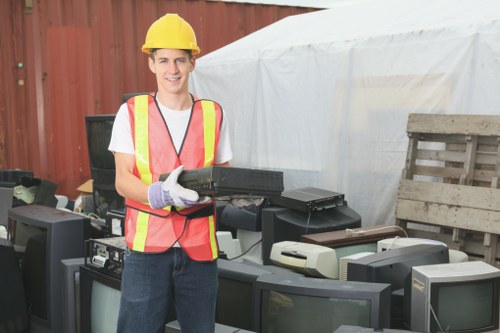 A young male worker wearing a yellow safety helmet, high-visibility vest with orange and yellow stripes, and purple work gloves is standing outdoors in front of a collection of discarded electronic rubbish, including several old computer monitors and printers, arranged on the ground. The background shows a red metal fence and a white tent-like structure, indicating a waste collection or disposal site, possibly in Barking or nearby in East London. The worker is smiling and holding a black laptop, which he appears to be preparing to load for rubbish removal. The environment is well-lit with natural daylight, and the scene emphasizes the collection and management of electronic waste as part of commercial waste services offered by Commercial Waste Barking, focusing on professional rubbish removal in the local area.