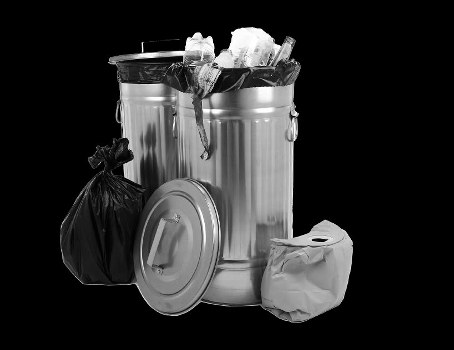 A large cylindrical metal rubbish bin with a fitted lid, positioned on a dark background. The bin contains visible waste materials such as crumpled paper and plastic packaging, with some items hanging over the edge and protruding from the top. To the left of the bin, there is a black plastic garbage bag, tightly tied at the top, leaning against the side. To the right of the bin, a crumpled paper bag is resting on the ground, and the lid of the bin, which appears to be made of metal or plastic, is placed on the ground in front of the bin, slightly tilted to the side. This scene is indicative of waste disposal, likely representing rubbish collection services available in Barking or the surrounding areas. The metallic surface of the bin has a reflective finish, contrasting with the dark background, and the overall composition focuses on waste management containers and discarded materials.