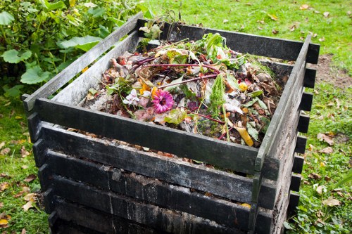 A large, rectangular black compost bin made of plastic with a slatted design, situated outdoors on a grassy area, filled with organic garden waste including colorful flower remnants, leafy greens, and plant stems. The contents are partially decomposed, showing various textures of plant material, some with damp, dark soil residue. Surrounding the bin are small green plants and grass, indicating a garden or backyard environment. The scene appears natural and well-lit, consistent with an outdoor setting in Barking, where commercial waste removal services by Commercial Waste Barking might assist with garden refuse disposal.