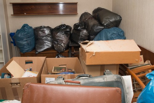 A cluttered indoor space with several black and blue plastic rubbish bags stacked on a wooden shelf against a beige wall, some partially covered with a large, slightly torn cardboard box. In the foreground, there are open cardboard boxes filled with miscellaneous items, including papers and small objects, placed on a surface or floor. A large, gray industrial vacuum cleaner or waste disposal unit is partially visible near the lower edge, with the scene illuminated by indoor lighting. The environment appears to be a storage or utility room, and the overall impression indicates it is an area cleared of waste as part of rubbish removal, possibly conducted by a company such as Commercial Waste Barking, serving the Barking area and postcode region.