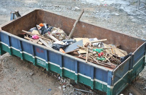 A large rectangular construction skip positioned on a gravel and dirt surface, filled with mixed waste materials including wooden planks, broken pieces of drywall or plasterboard, black garbage bags, and plastic packaging. The skip is made of metal with a rust-colored interior and a blue exterior featuring green and white horizontal stripes. The surrounding environment suggests an outdoor area, possibly a construction or renovation site, with uneven ground and loose gravel. Natural daylight illuminates the scene, highlighting the texture of the waste and the rough surface beneath. The skip is situated in a way that indicates it is intended for rubbish removal, with no other objects or equipment visible in the immediate background. Commercial Waste Barking services this area, facilitating the proper disposal of construction or renovation debris in the local postcode area.