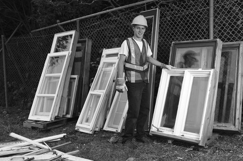 A male worker wearing a white safety helmet, high-visibility vest, and dark trousers stands outdoors on a patch of grass adjacent to a chain-link fence, next to a collection of discarded white window frames with glass panes. The frames are leaning against the fence and arranged in a row, with some stacked vertically and others resting horizontally on the ground. The window frames vary in size and are made of materials such as timber or UPVC, featuring smooth finishes and visible glass panels. The surrounding environment suggests a site for waste collection or disposal, with additional loose window components and wooden offcuts scattered at the worker's feet. The lighting appears natural, likely daytime, contributing to a clear view of the scene, which aligns with services offered by Commercial Waste Barking for rubbish removal and waste management in the local area. This image emphasizes the process of waste collection, including the handling of building or renovation debris, pertinent to commercial waste services in Barking or nearby postcode districts.
