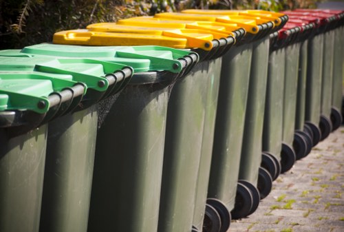 A row of green wheeled rubbish bins with yellow, green, and red lids, lined up along a paved pathway on the edge of a driveway or street surface. The bins are made of durable plastic with black rubber wheels and black handles, and they are positioned side by side, slightly angled downward, indicating they are ready for collection or disposal. The background features a hint of foliage and outdoor greenery, suggesting a residential or commercial area in Barking. The scene is captured in natural daylight, which highlights the clean, smooth surface of the bins and the vibrant color-coding of the lids, reflecting typical waste management practices employed by companies such as Commercial Waste Barking in the local area for rubbish collection services.