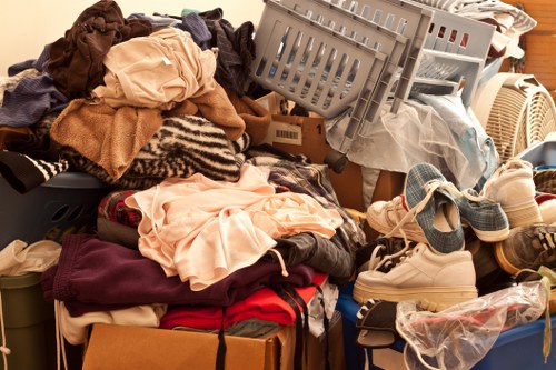 A cluttered assortment of domestic clothing and footwear piled on top of cardboard boxes and plastic crates, with some garments including shirts, sweaters, and trousers visibly folded or draped, alongside pairs of shoes and sandals scattered across the scene. The items appear to be a mix of clean and used, with fabrics of various textures such as cotton, wool, and synthetic materials, in different colours including beige, maroon, and light blue. The setting appears to be an indoor space, possibly a storage area or a room designated for clearing out household items, with a background that suggests a domestic environment. The overall arrangement indicates an accumulation of rubbish or unwanted personal belongings awaiting removal or disposal, aligning with the services offered by Commercial Waste Barking for efficient rubbish removal in the Barking area.